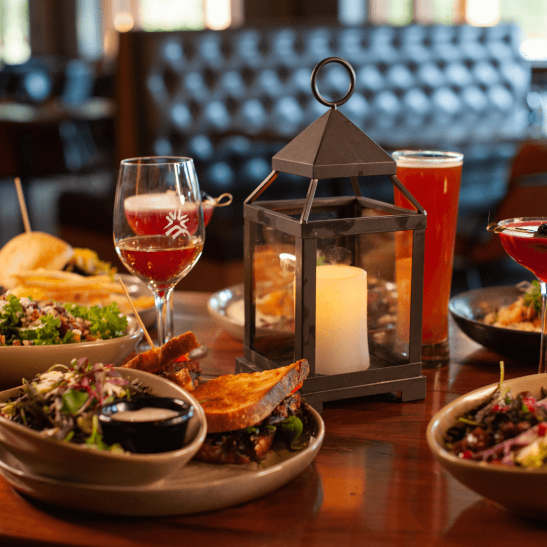 A table with various dishes and drinks, including sandwiches, salads, cocktails, and a pint of beer, centered around a decorative lantern with a lit candle.