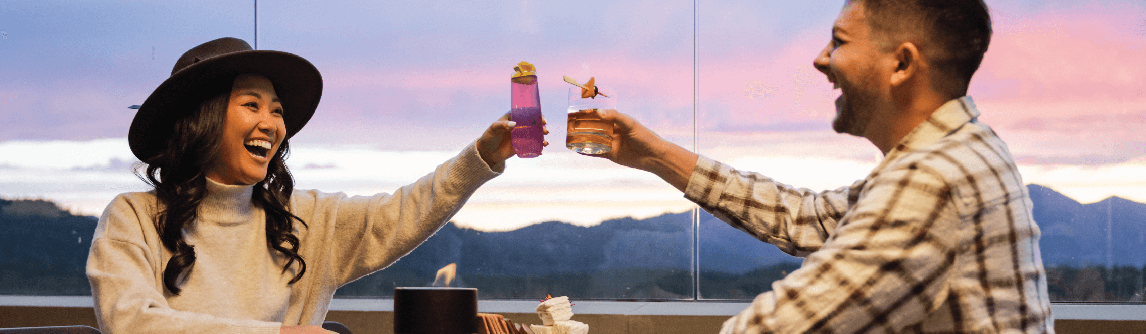 Two people sitting at an outdoor table, smiling and clinking glasses at sunset. The woman wears a hat; the man wears a plaid shirt. Mountains and sky are visible in the background.