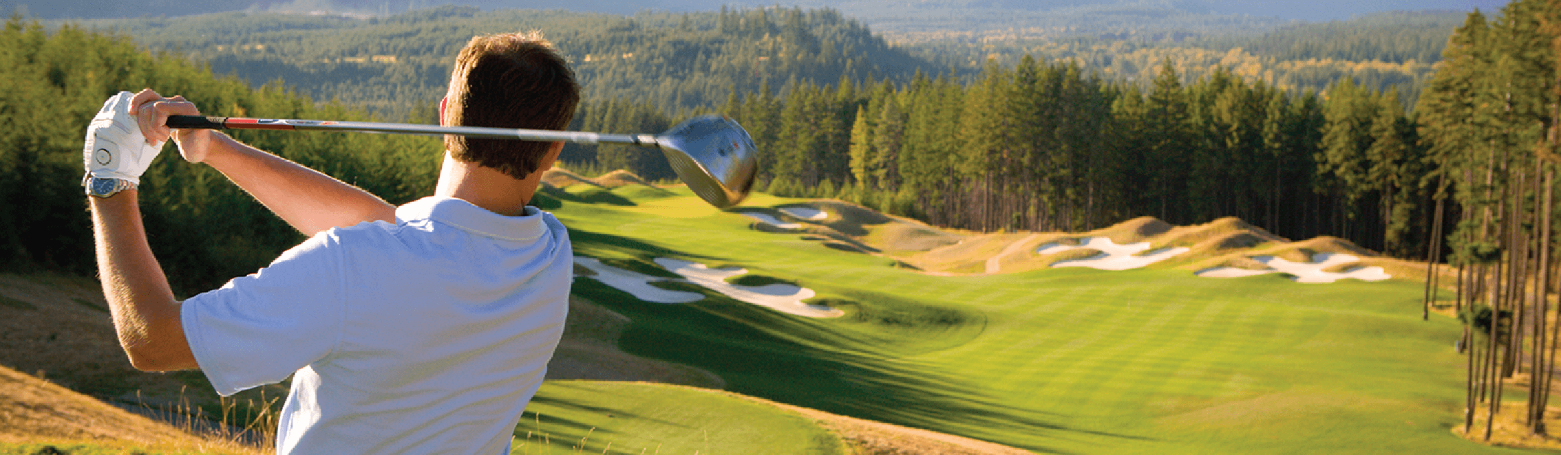 A golfer swings a club on a lush golf course, surrounded by trees and hills, under a clear sky.