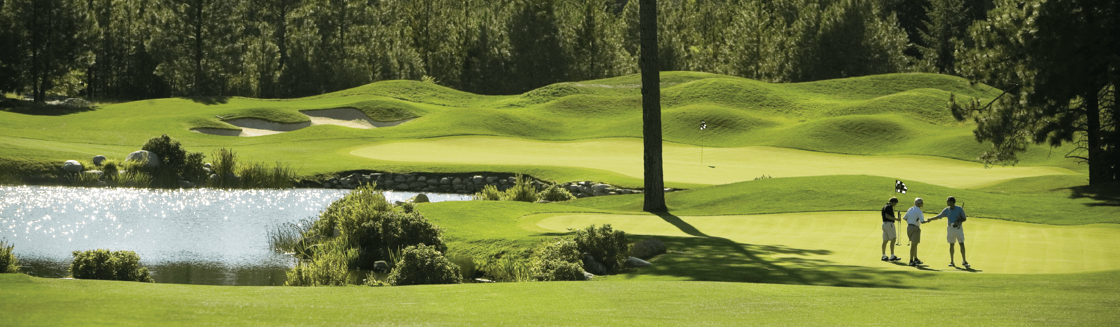 Three people standing on a golf course near a pond, surrounded by trees and sand traps, with one person holding a golf club.