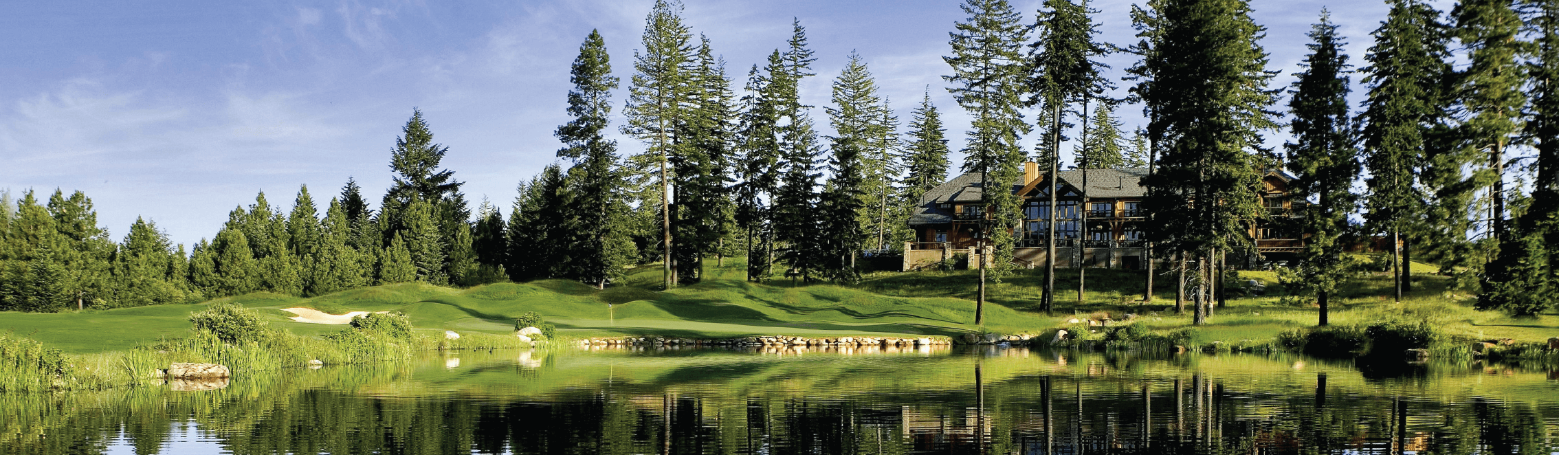 A large house surrounded by tall trees beside a serene lake, with a manicured lawn and clear blue sky.