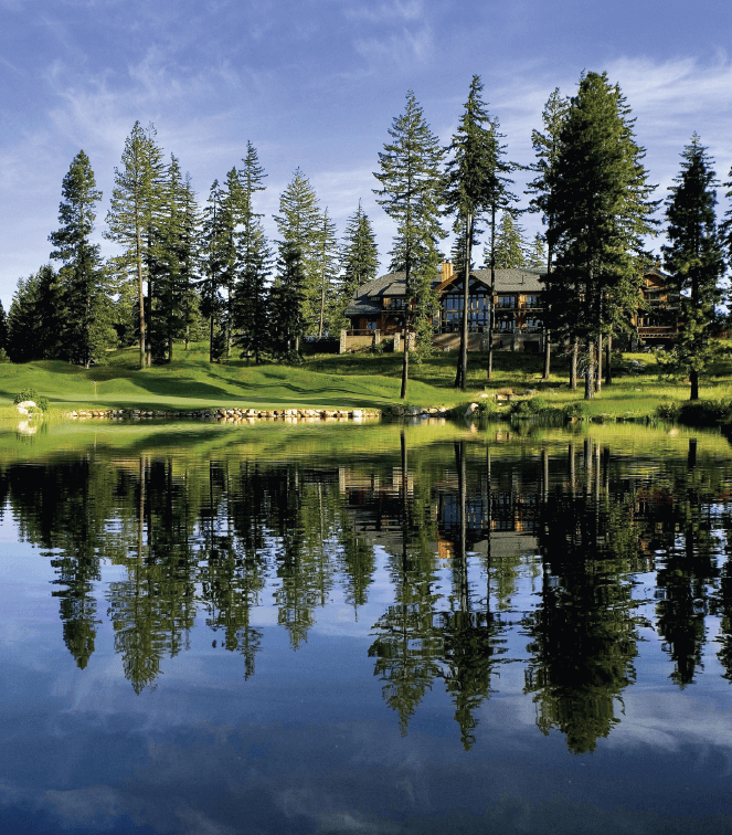 Lakeside view of a lodge surrounded by pine trees, with clear water reflecting the trees and building under a blue sky.