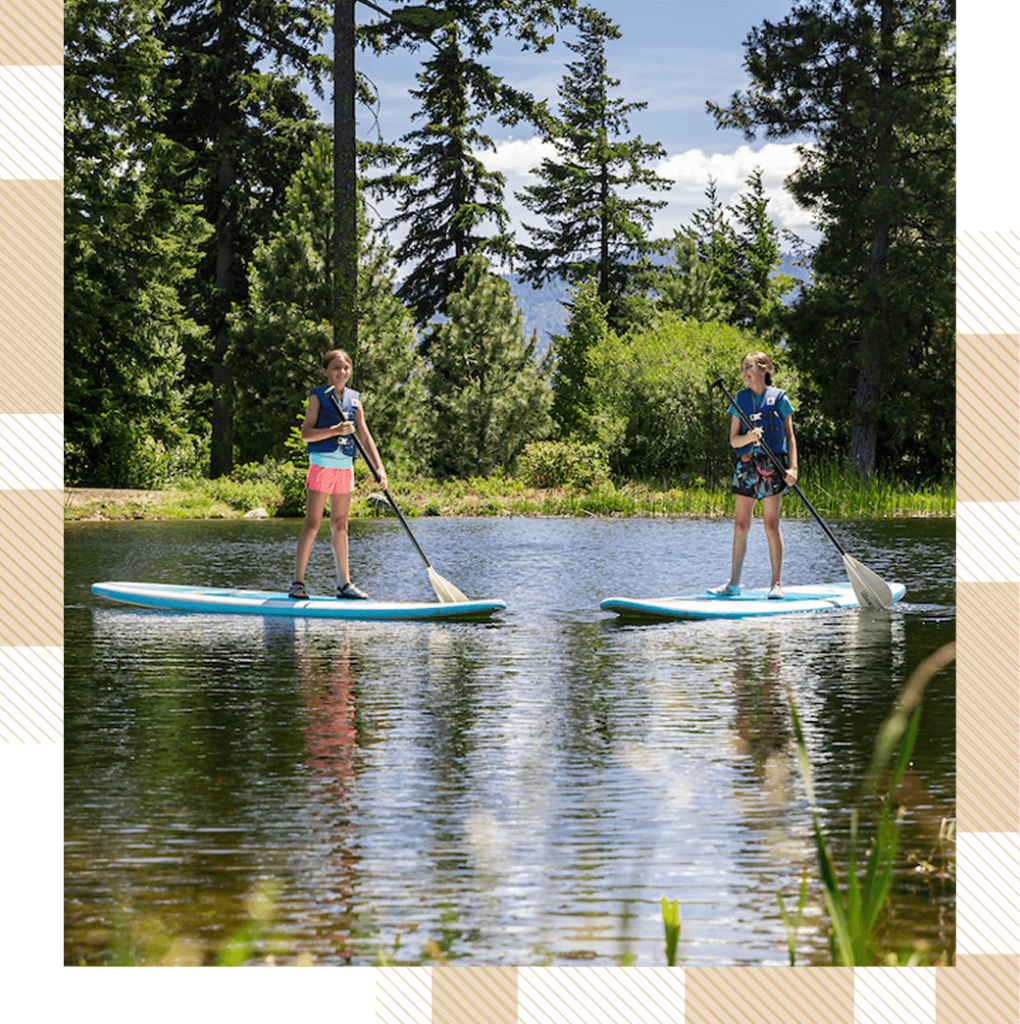 Two people paddleboarding on a calm lake, surrounded by trees and clear skies.