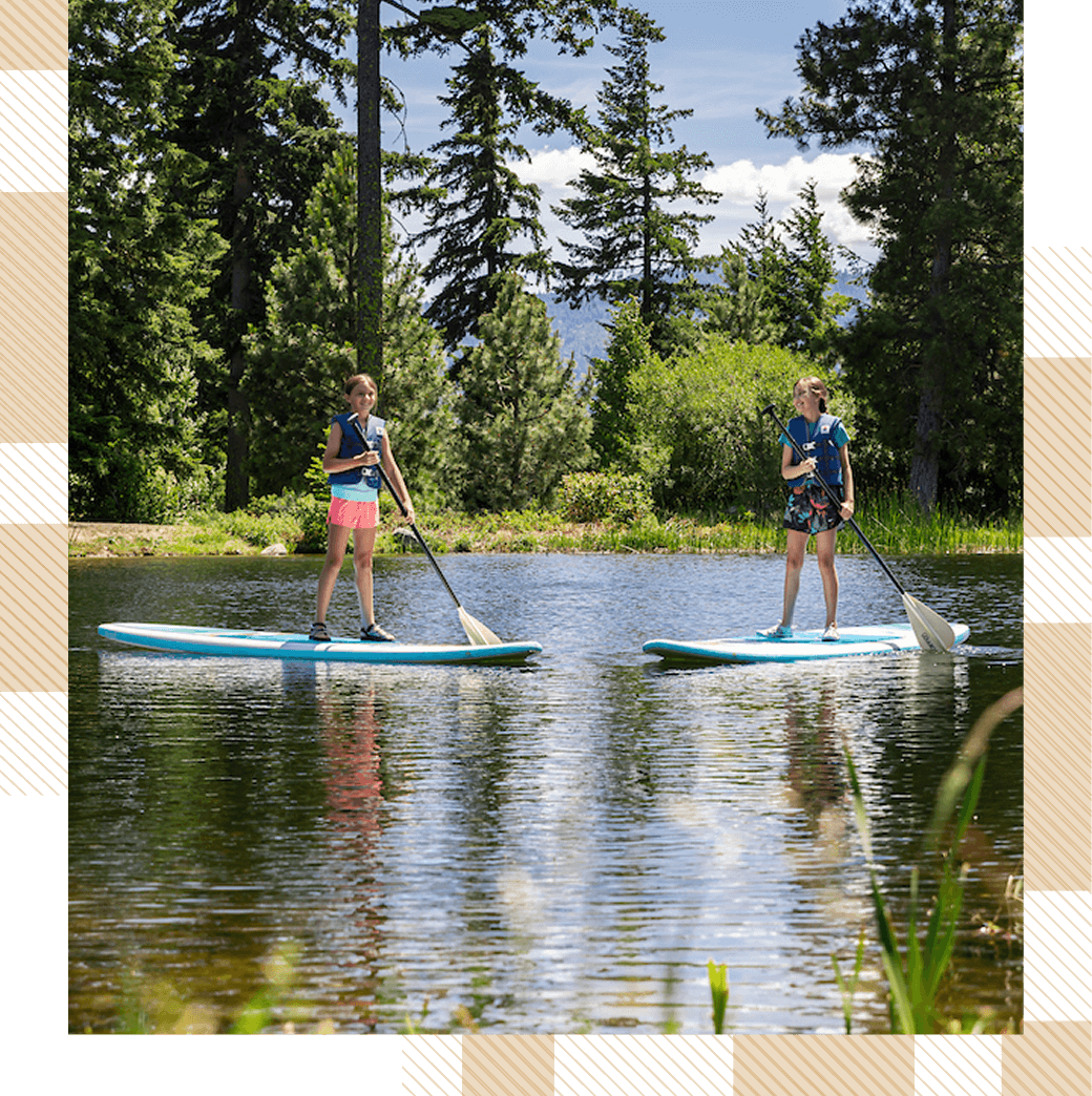 Two people paddleboarding on a calm lake, surrounded by trees and clear skies.