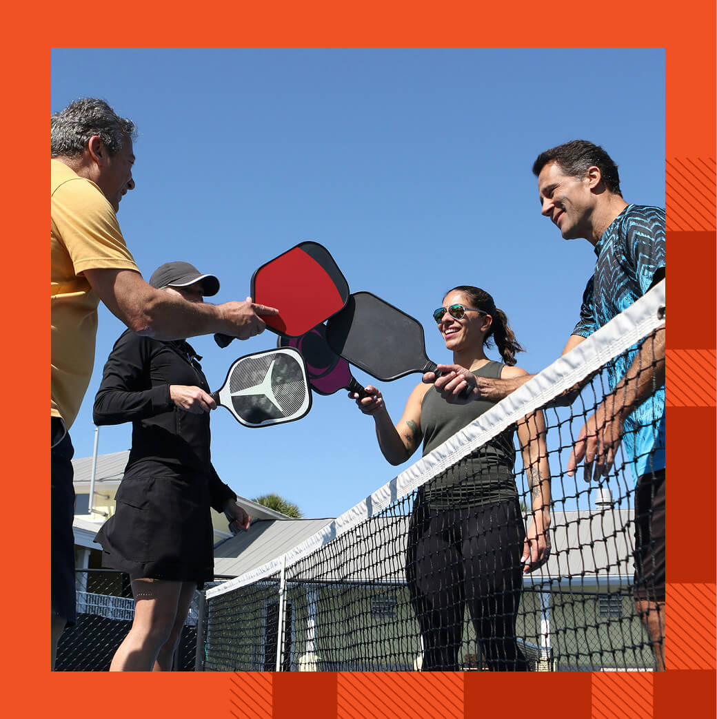 Four people holding paddles over a net on a pickleball court, with clear skies and a building in the background.