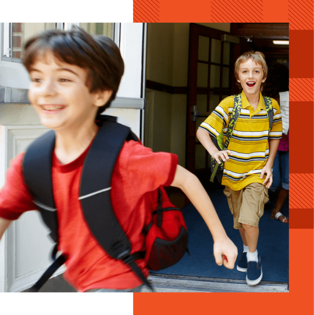 Two boys with backpacks are running out of a school building, smiling and appearing excited.