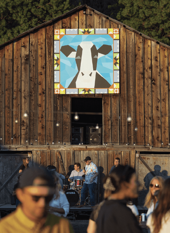A band performs in front of a rustic wooden barn with a large, geometric cow mural above them; people gather in the foreground.