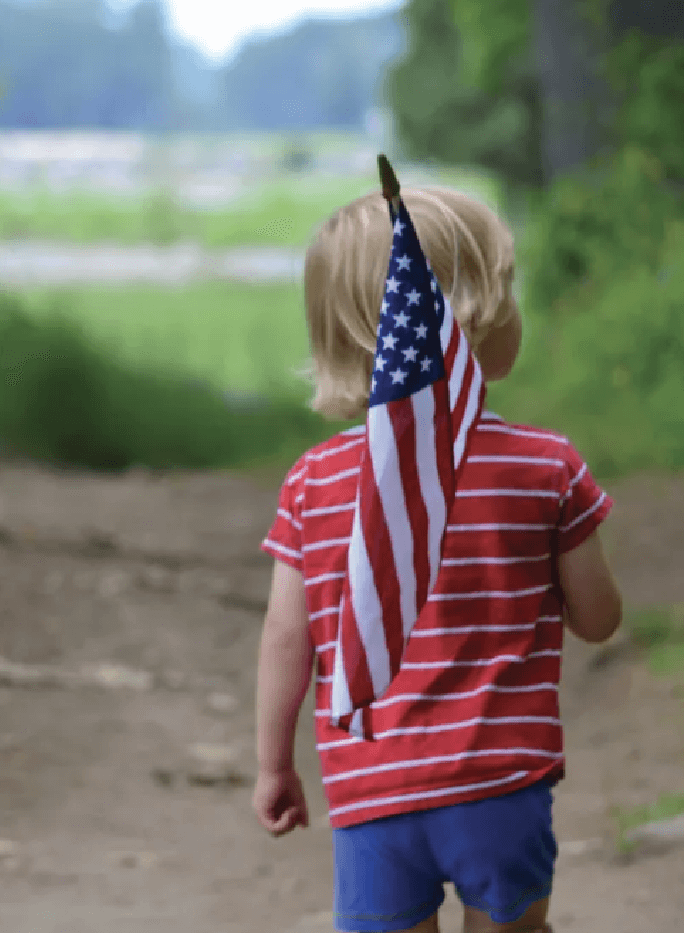 A young child wearing a red striped shirt and blue shorts walks outdoors holding an American flag over their shoulder.