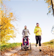 A man and woman jog outdoors in autumn, with the man pushing a child in a stroller along a tree-lined path covered in fallen leaves.