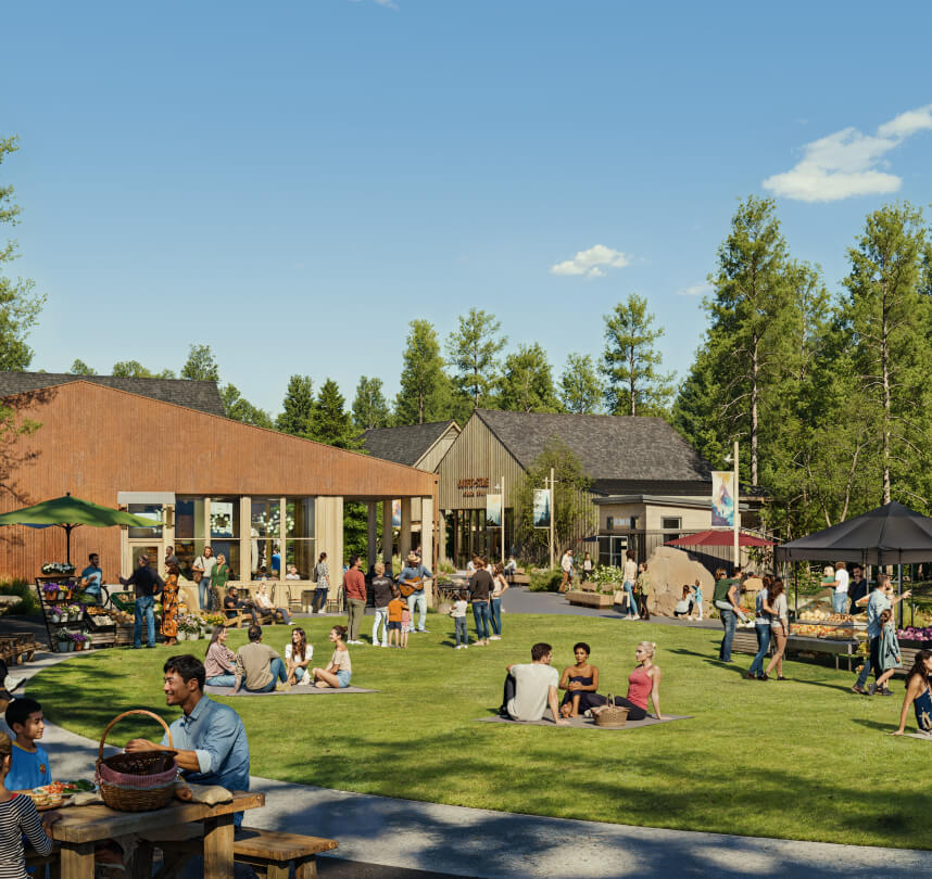 People gather and relax on a grassy lawn outside modern buildings on a sunny day, with groups sitting on the grass and at picnic tables. Trees and blue sky are in the background.