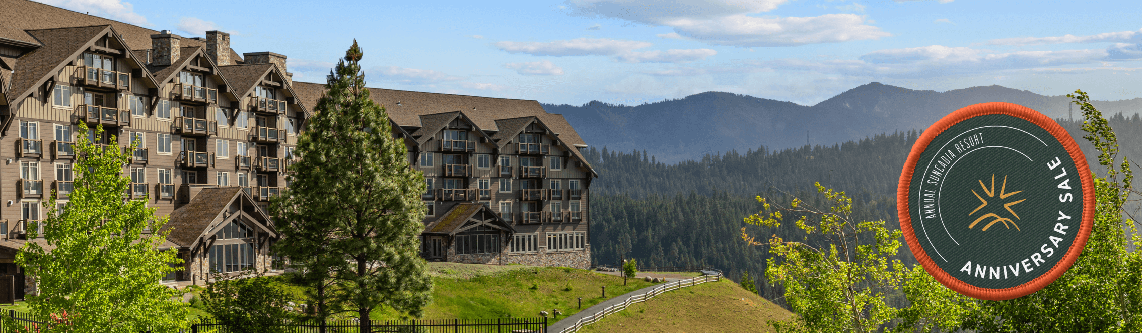 A large lodge-style hotel sits on a green hillside with pine trees, overlooking forested mountains under a blue sky. A circular badge on the right reads Anniversary Sale.