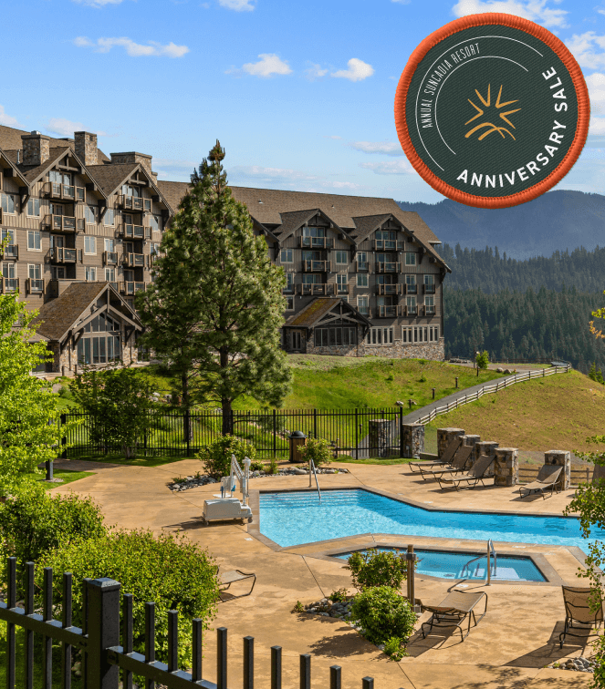 A mountain resort with rustic lodge-style buildings overlooks a scenic valley. A fenced outdoor pool area with lounge chairs sits in the foreground. An Anniversary Sale badge is in the upper right corner.