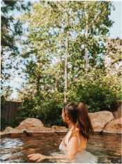 A woman with brown hair in a ponytail sits in a natural-looking outdoor pool, surrounded by rocks, greenery, and tall trees under a clear sky.