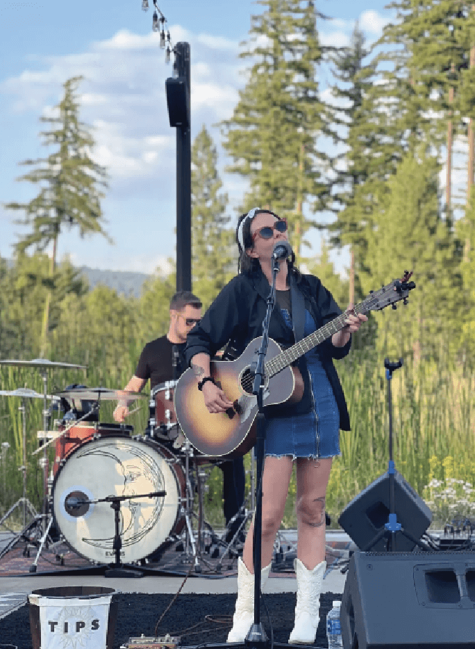 A woman plays guitar and sings into a microphone on an outdoor stage, with a drummer performing behind her. Trees and a clear sky are visible in the background.