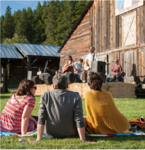 Four people sit on the grass facing a small outdoor stage where a band is performing in front of a rustic wooden building.