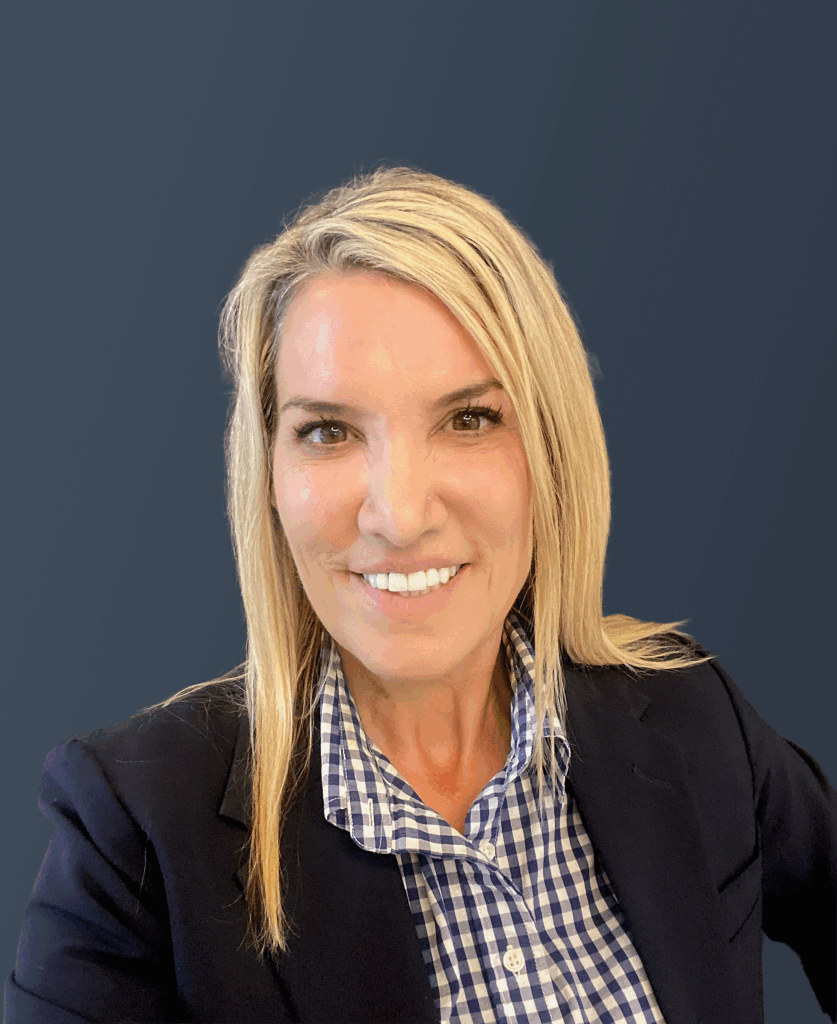 A woman with straight blonde hair, wearing a navy blazer over a blue and white checkered shirt, smiles at the camera against a plain dark background.