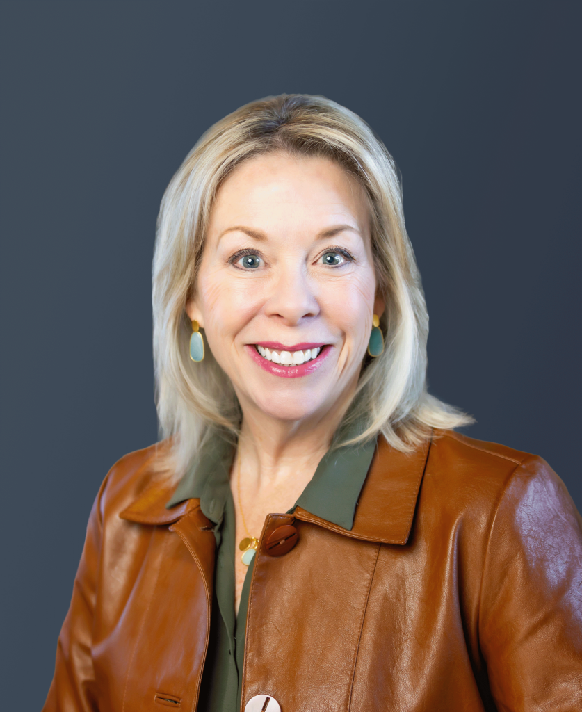 Smiling woman with blonde hair wearing a brown leather jacket and green shirt, posed against a plain dark background.