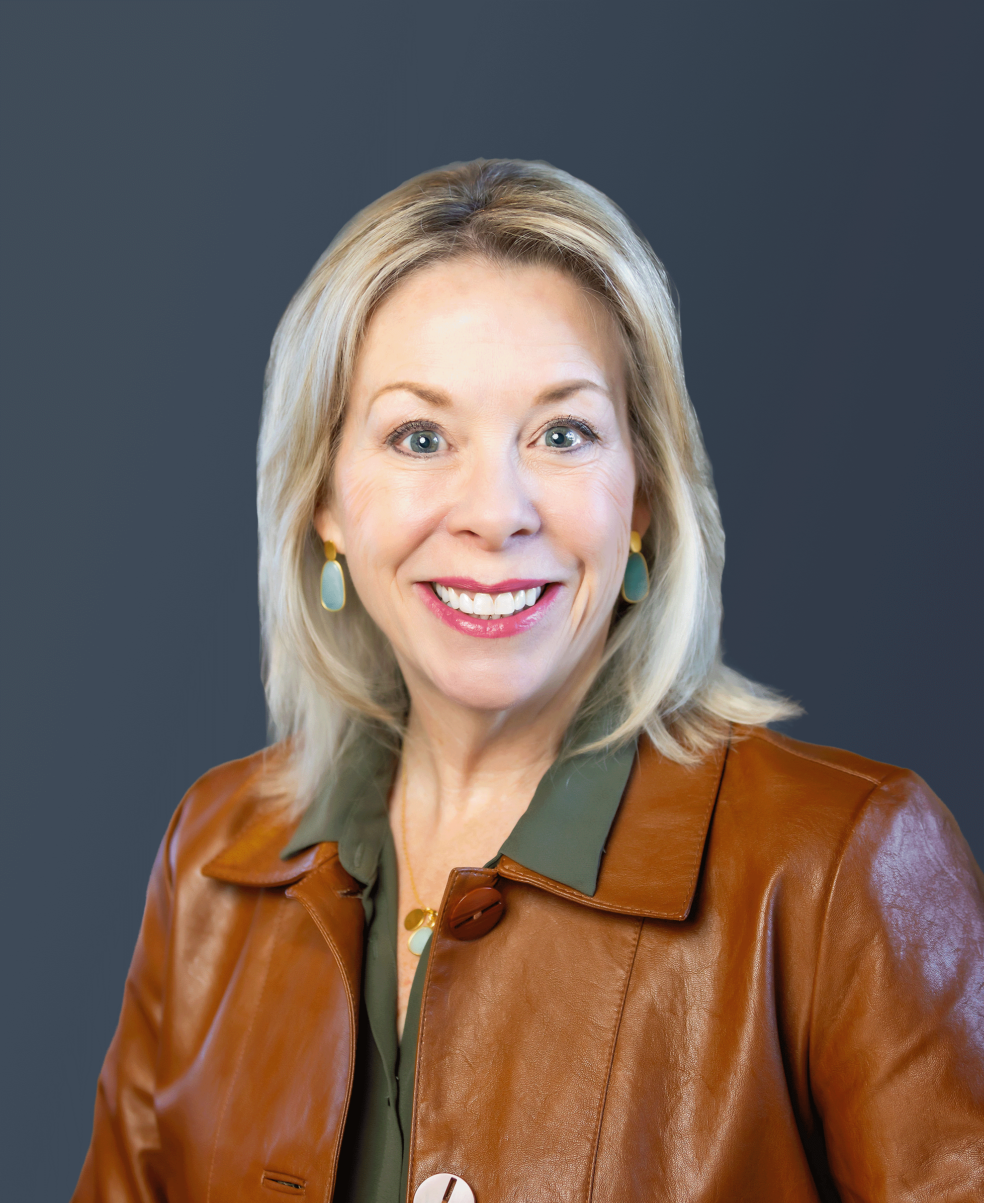 Smiling woman with blonde hair wearing a brown leather jacket and green shirt, posed against a plain dark background.