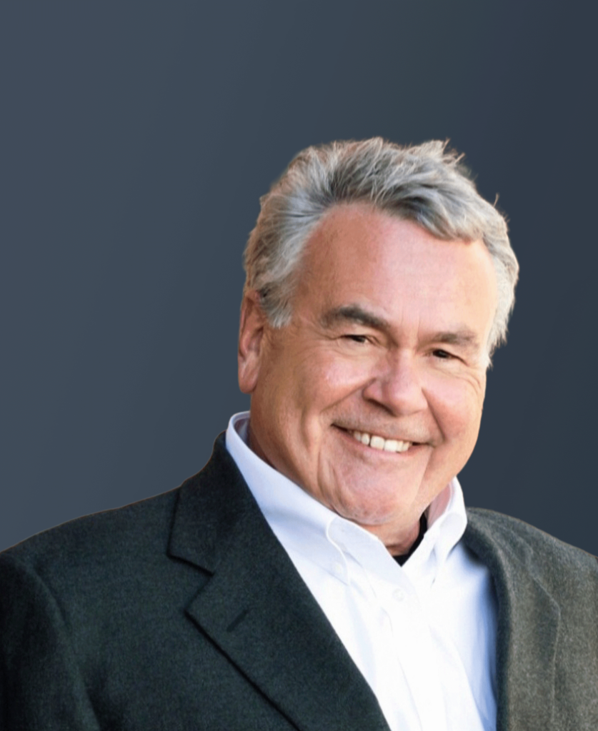 A smiling older man with gray hair wearing a dark blazer and white collared shirt, posed in front of a plain, dark background.