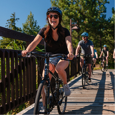 A group of people wearing helmets ride bicycles across a wooden bridge on a sunny day, surrounded by trees and greenery.