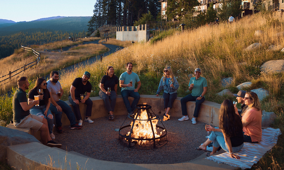 A group of people sit around a fire pit outdoors on a grassy hillside, holding drinks and talking as the sun sets.