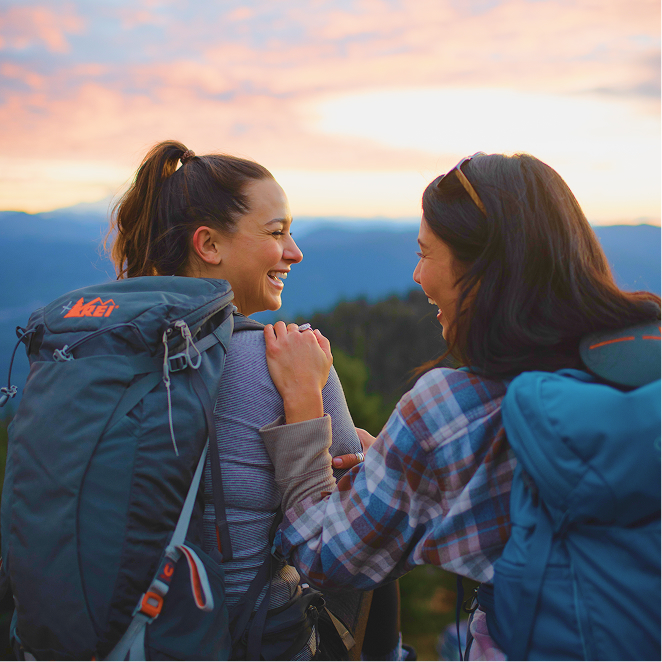 Two people with backpacks smile at each other outdoors, with mountains and a colorful sunset sky in the background.