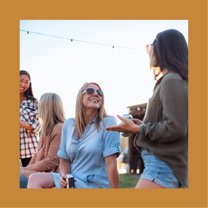 Four women socialize outdoors in casual clothing on a sunny day; one woman in a blue dress is smiling and holding a drink can.