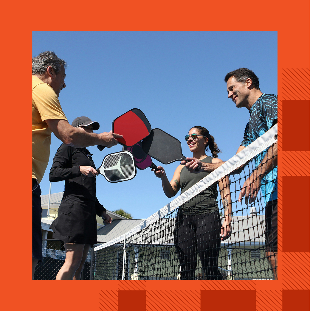 Four people stand at a pickleball net, smiling and tapping paddles together on a sunny day.