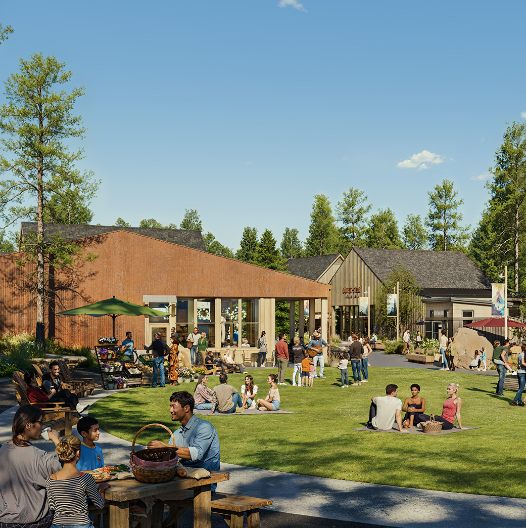 People are gathered outdoors on a grassy lawn near modern buildings, sitting on benches and blankets, socializing and enjoying a sunny day under a clear blue sky.