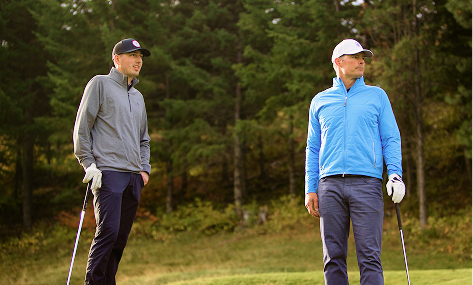Two men wearing jackets, hats, and golf gloves stand on a golf course, each holding a golf club, with trees in the background.