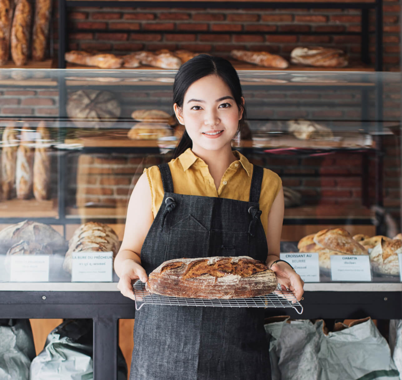 A woman wearing an apron stands in a bakery, holding a loaf of bread on a cooling rack, with shelves of assorted bread loaves in the background.