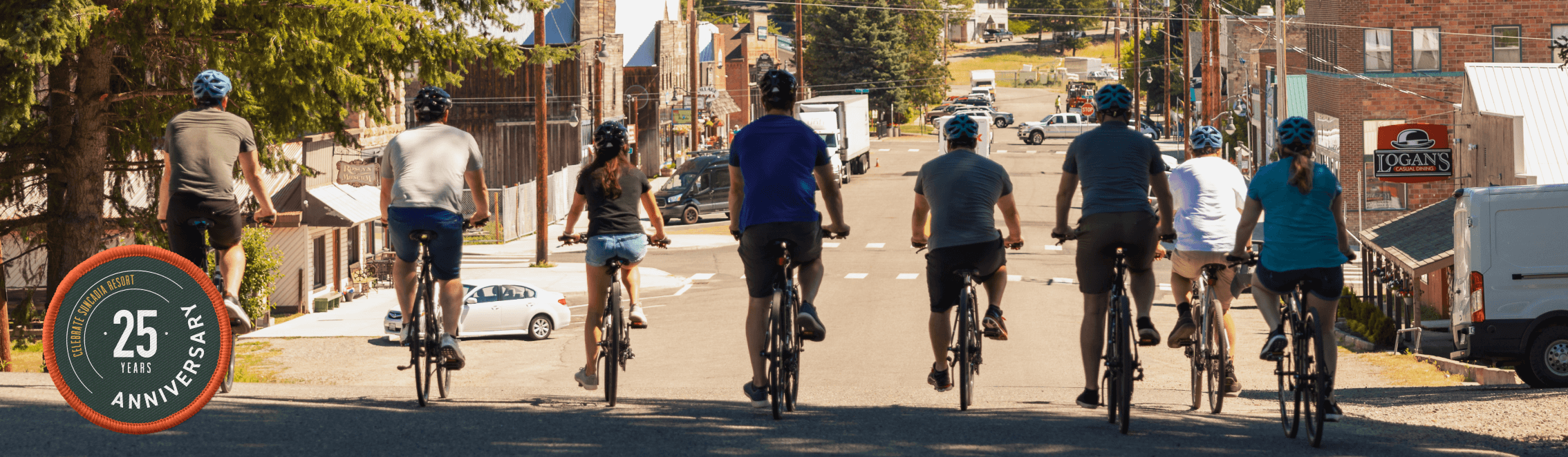 Eight people wearing helmets ride bicycles down a street in a small town, with a patch on the left marking a 25-year anniversary.