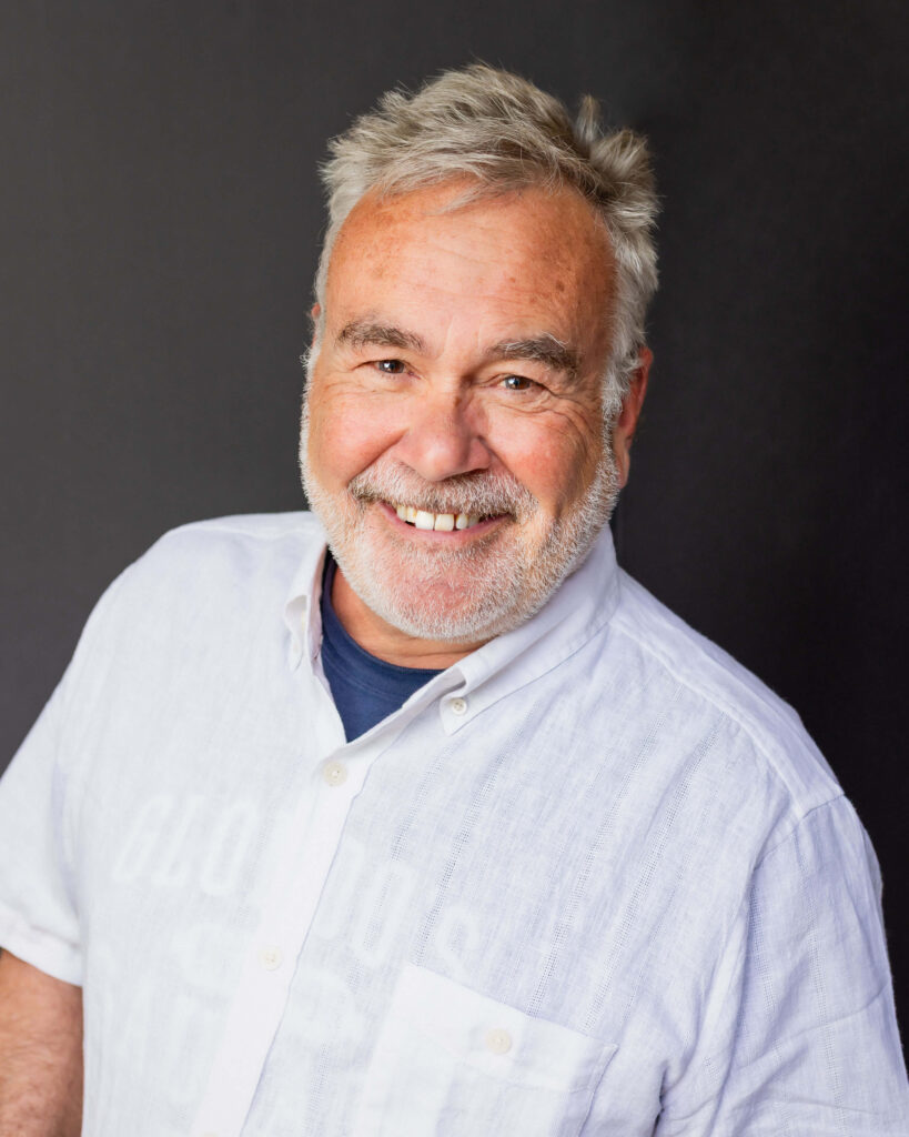 An older man with gray hair and a beard smiles at the camera, wearing a white button-up shirt against a plain dark background.