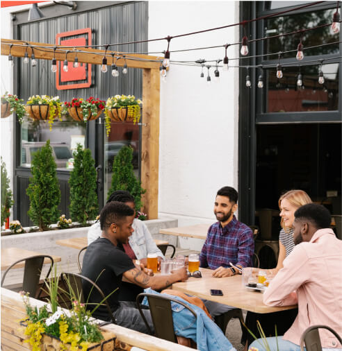 Five people sit around an outdoor table at a restaurant, drinking beer and talking. String lights and plants decorate the patio area.