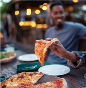 A person holds a slice of cheese pizza over a wooden table with a whole pizza, empty plates, and a smiling person in the background.