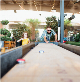 A man and a young girl play shuffleboard together at an outdoor venue with trees and string lights in the background.