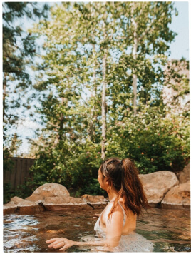 A person with long brown hair in a ponytail sits in a natural outdoor pool, surrounded by rocks and trees, looking away from the camera.