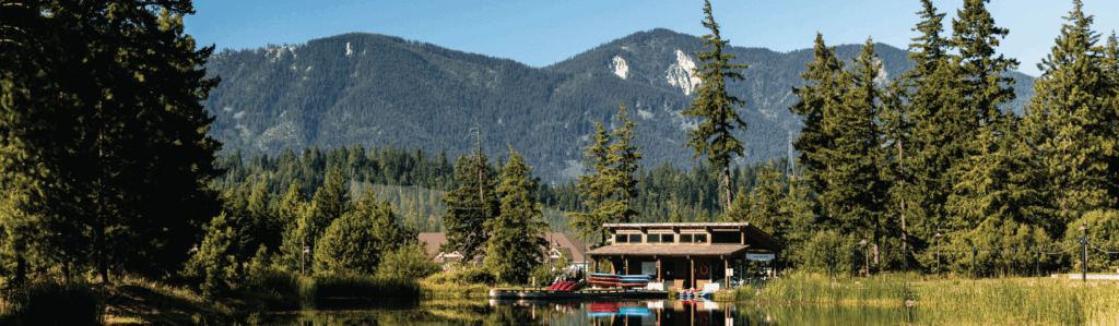 A wooden boathouse with stacked kayaks sits by a calm lake, surrounded by tall trees and mountains under a clear blue sky.