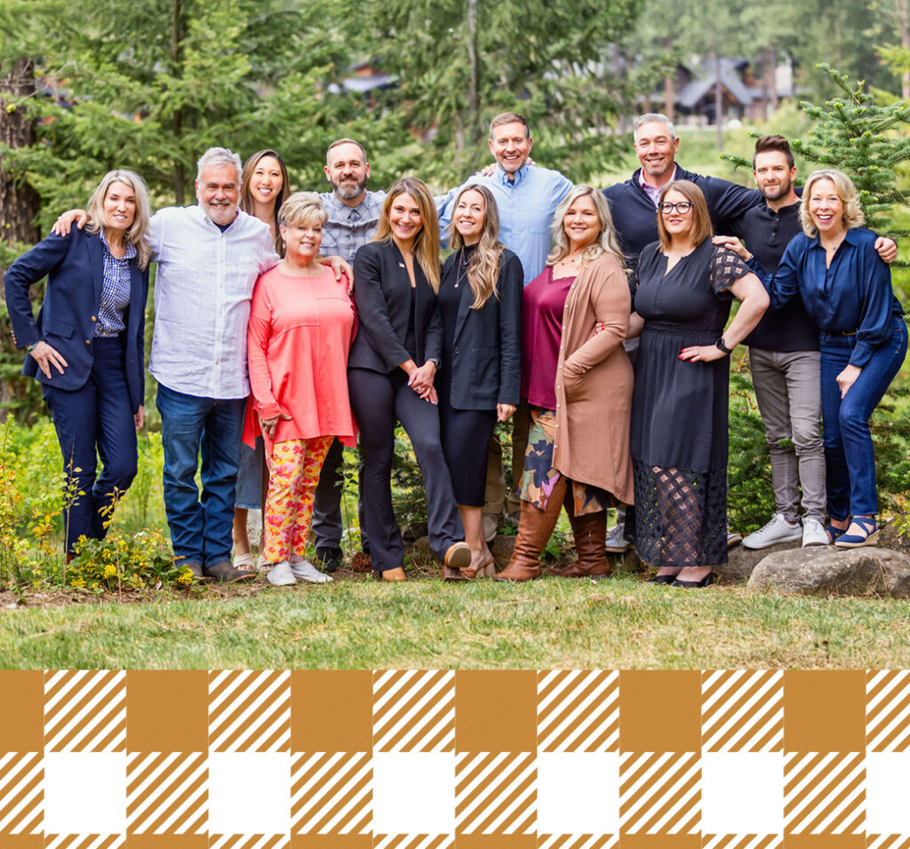 A group of fourteen adults poses together outdoors in front of trees, standing on grass and smiling at the camera.