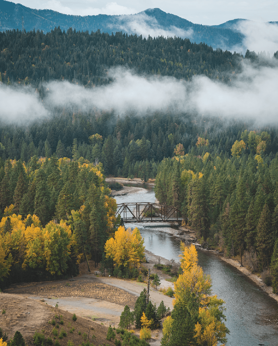 A river winds through a forested landscape with autumn foliage, featuring a metal bridge and low-hanging clouds over distant mountains.