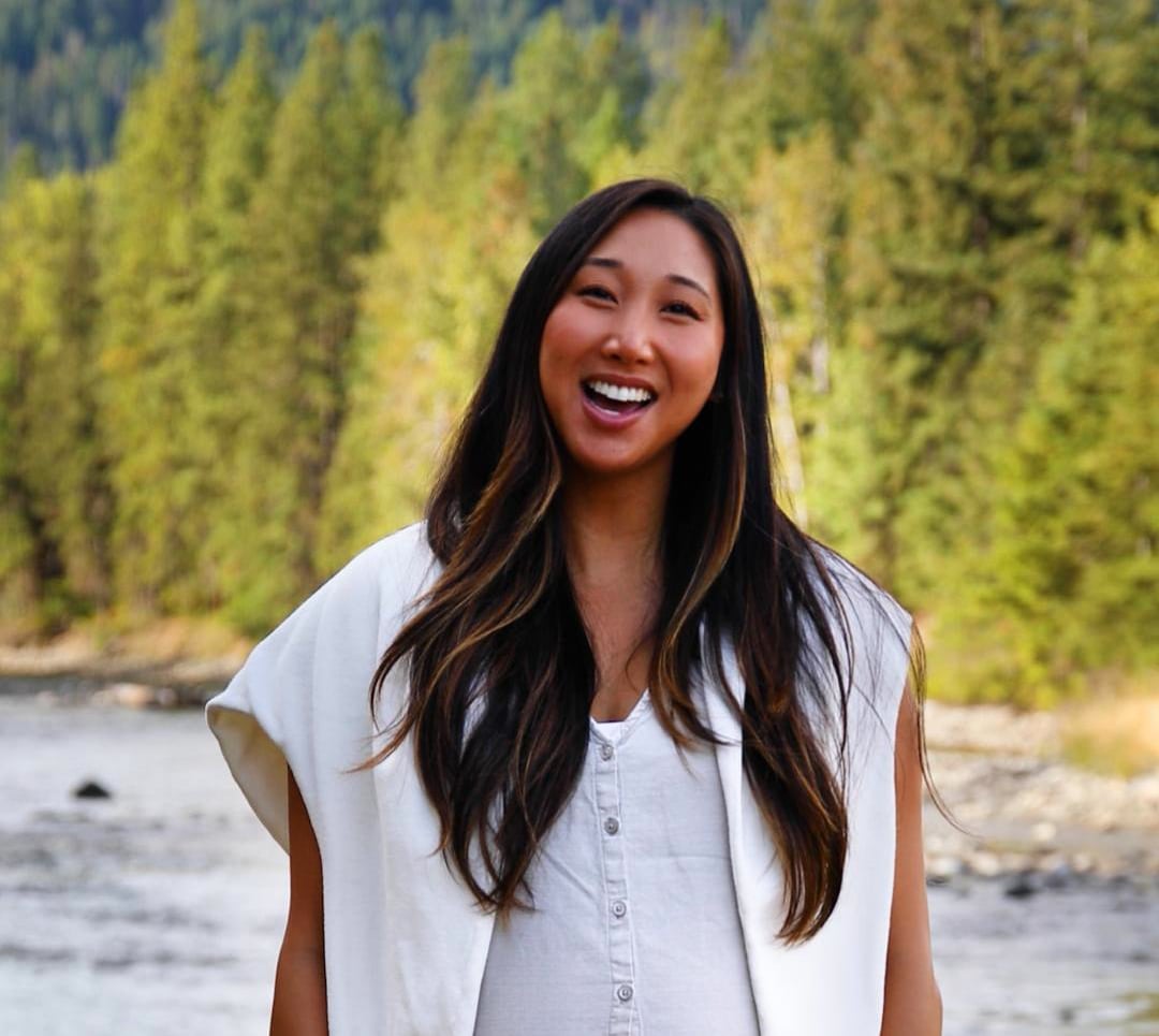 Woman with long dark hair wearing a white shirt and vest stands outdoors, smiling in front of a river and forested background.