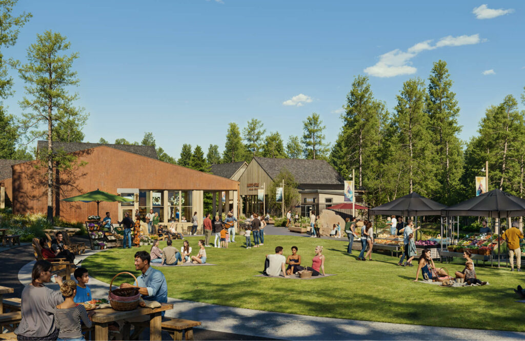 People gather and relax on a grassy lawn surrounded by trees, sitting at picnic tables and on the grass near market stalls and buildings on a sunny day.