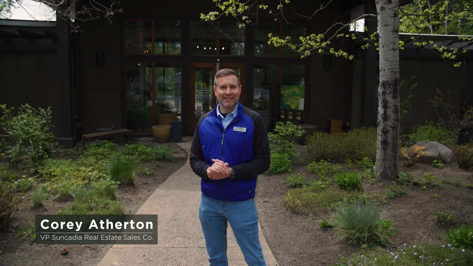 A man stands on a garden path in front of a modern building, wearing a blue vest and jeans. Text overlay reads "Corey Atherton, VP Suncadia Real Estate Sales Co.