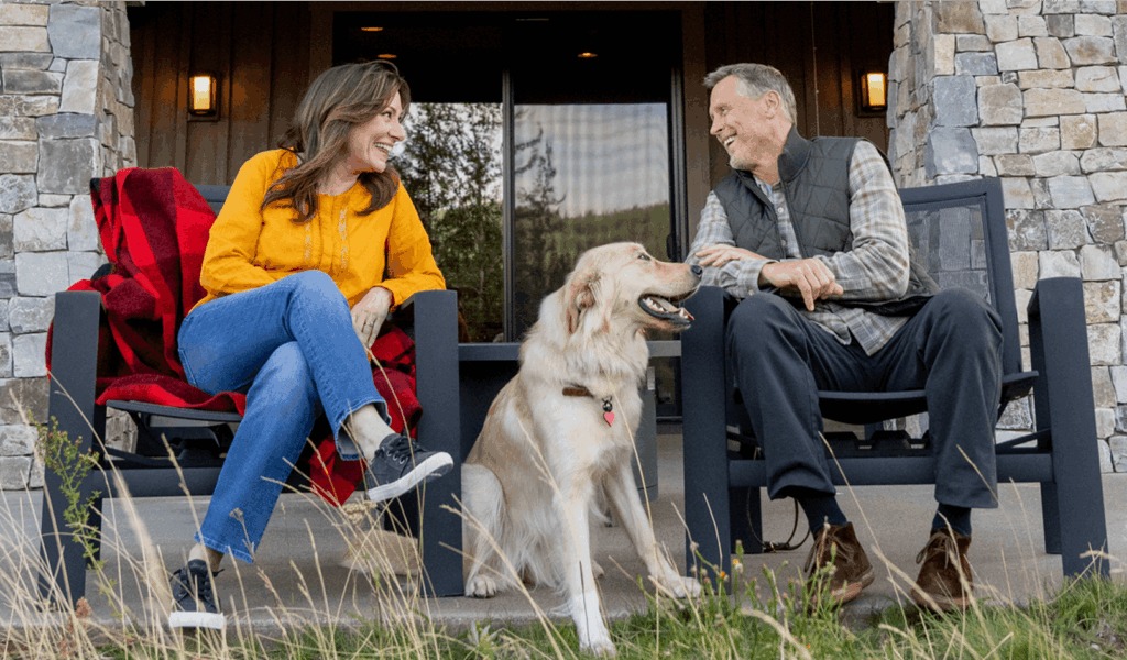 Two people sit on chairs on a porch, smiling at each other, with a golden retriever dog between them. Stone pillars and wooden walls are in the background.