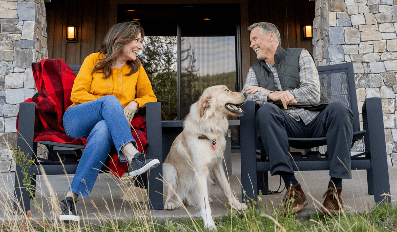 Two people sit on chairs on a porch, smiling at each other, with a golden retriever dog between them. Stone pillars and wooden walls are in the background.