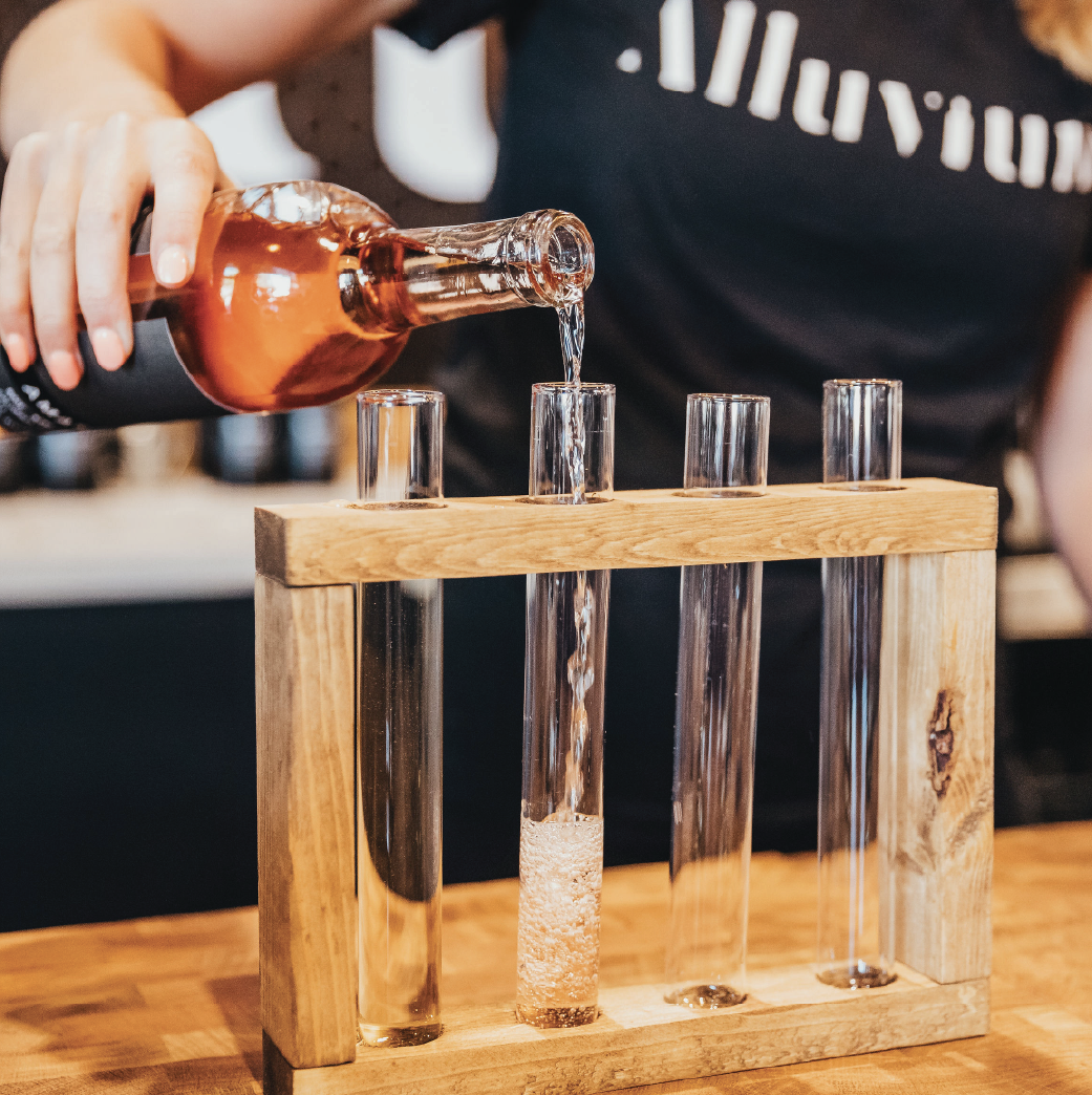 A person pours liquid from a bottle into a test tube held in a wooden rack with three other empty test tubes.