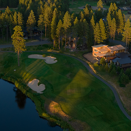 Aerial view of a golf course with sand traps, a water hazard, surrounding trees, and houses near the green in the evening sunlight.