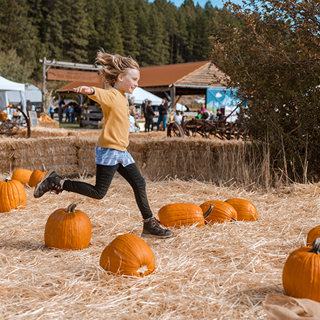 A child runs and leaps over pumpkins arranged on straw at an outdoor fall festival, with tents and trees in the background.