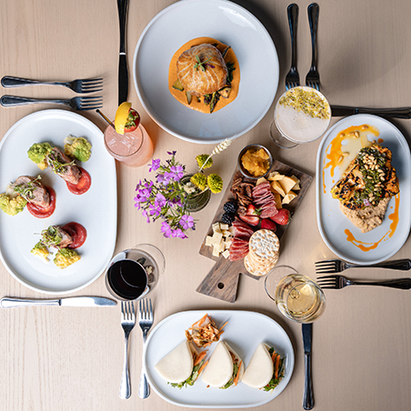 Overhead view of a table set with various plated dishes, charcuterie board, drinks, and cutlery, with a small floral centerpiece.