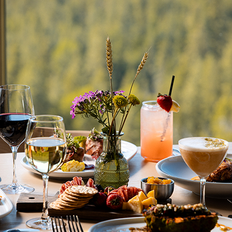 A table set with wine, cocktails, assorted appetizers, crackers, cheese, fruit, and a vase of wildflowers, with a forest view in the background.